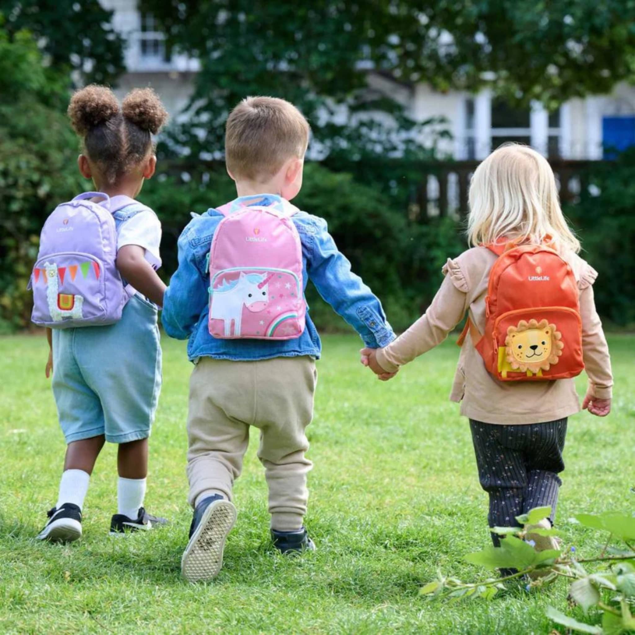 Drei Kinder mit farbenfrohen Kinderrucksäcken beim Spaziergang auf einer grünen Wiese