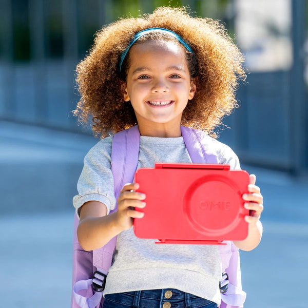Kind hält Lunchbox mit Thermosbehälter OmieBox Up – Cherry Pink draußen vor moderner Fassade